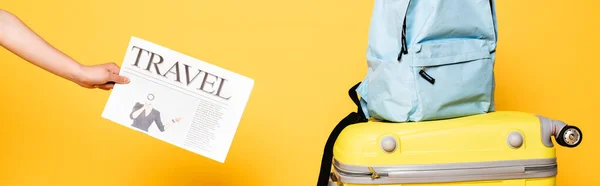 Cropped view of woman holding travel newspaper near blue backpack on travel bag on yellow, panoramic shot — Stock Photo