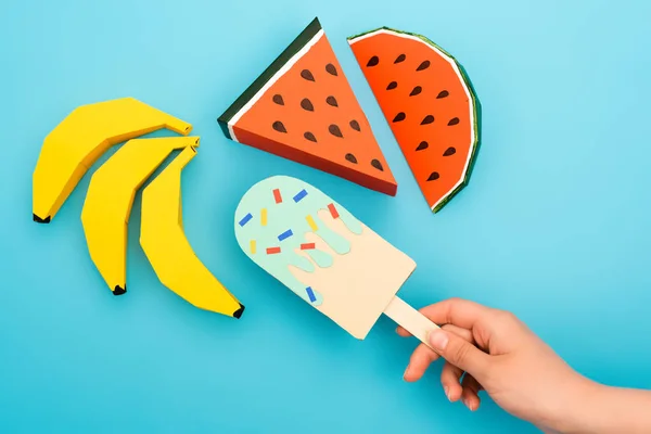 Cropped view of woman holding paper ice cream with sprinkles near watermelon, bananas on blue background — Stock Photo