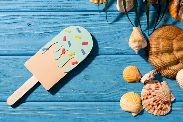 Верхний вид бумажного мороженого с брызгами рядом с раковинами и пальмовым листом на деревянном синем фоне — стоковое фото