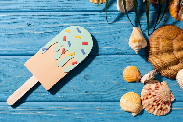 Vue du dessus de la crème glacée en papier avec des aspersions près des coquillages et de la feuille de palme sur fond bleu bois — Photo de stock