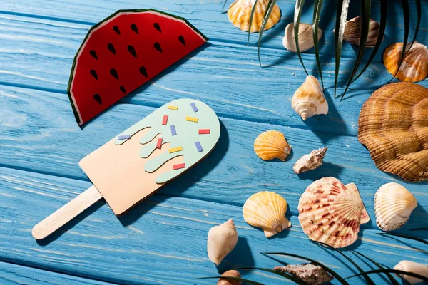 Vue du dessus de la crème glacée en papier avec des aspersions et de la pastèque près des coquillages et des feuilles de palmier sur fond bleu bois — Photo de stock