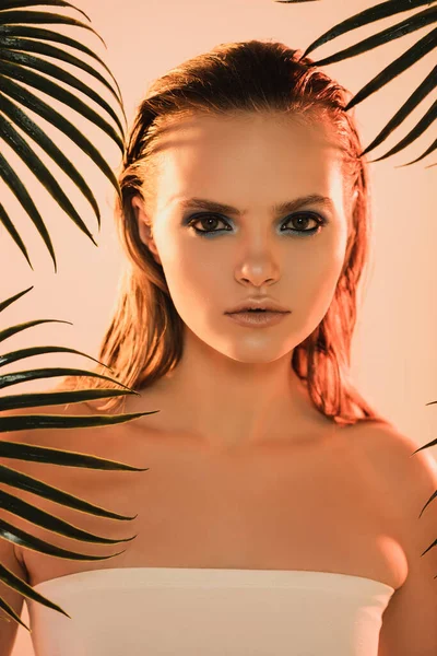 Beautiful woman with makeup looking at camera near palm leaves on beige — Stock Photo