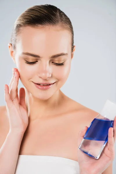 Glückliche Frau schaut Flasche mit Toner an und berührt Gesicht isoliert auf grau — Stockfoto