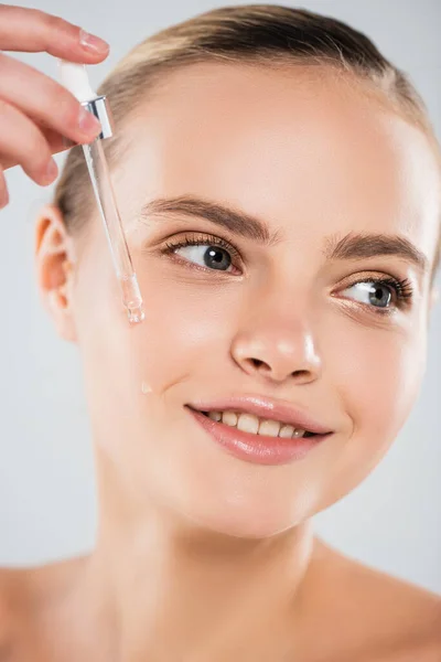 Nahaufnahme einer glücklichen Frau, die eine Pipette hält und Serum auf grau isoliert aufträgt — Stockfoto