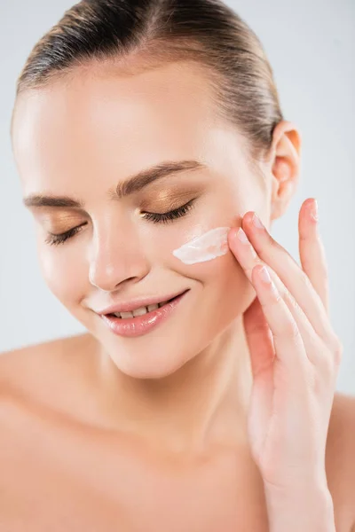 Mujer alegre con los ojos cerrados aplicando crema facial aislada en gris - foto de stock