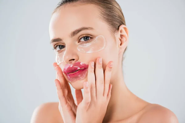 Jolie femme avec masque à lèvres et patchs pour les yeux touchant visage propre isolé sur gris — Photo de stock