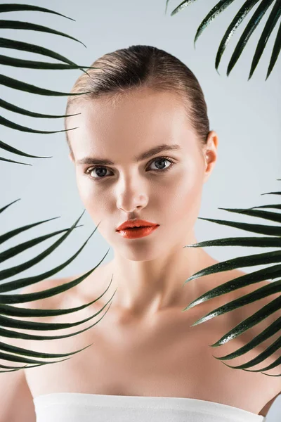 Pretty woman with makeup looking at camera near palm leaves on white — Stock Photo