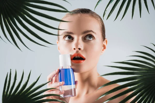 Mulher surpreso com maquiagem segurando garrafa com toner perto de folhas de palma no branco — Fotografia de Stock