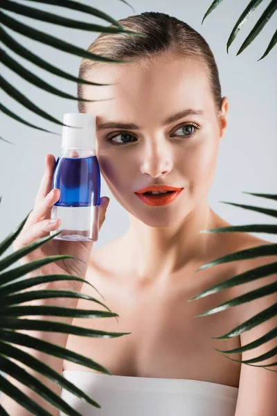 Mulher atraente com maquiagem segurando garrafa com toner perto de folhas de palma no branco — Fotografia de Stock