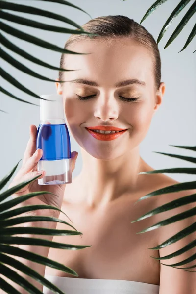 Femme heureuse avec bouteille de tenue de maquillage avec toner près des feuilles de palmier sur blanc — Photo de stock