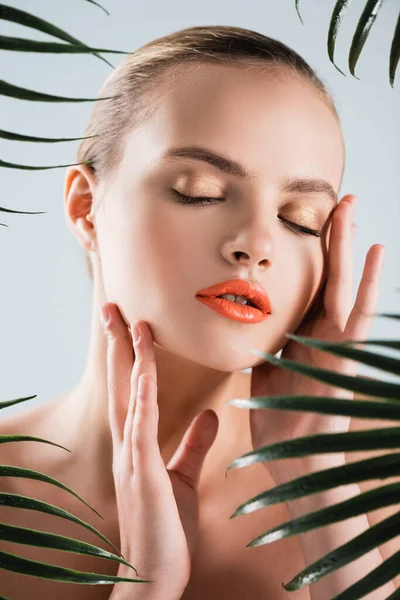 Foyer sélectif de la fille attrayante avec maquillage touchant visage près de feuilles de palmier sur blanc — Photo de stock