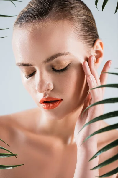 Attractive and naked girl with makeup touching face near palm leaves on white — Stock Photo