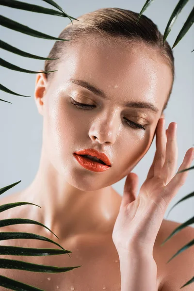 Donna attraente e bagnata con il trucco che tocca il viso vicino alle foglie di palma sul bianco — Foto stock