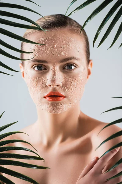 Mulher nua com maquiagem e esfregar no rosto olhando para a câmera perto de folhas de palma no branco — Fotografia de Stock