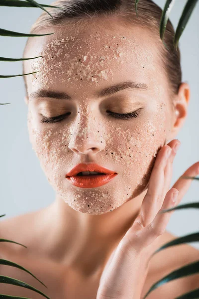 Enfoque selectivo de la mujer desnuda con maquillaje y exfoliante tocar la cara cerca de hojas de palma en blanco - foto de stock