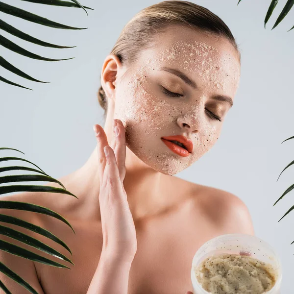 Naked woman applying scrub and holding container near palm leaves on white — Stock Photo