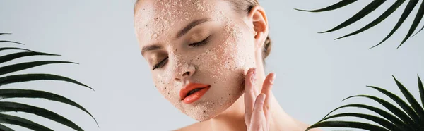 Panoramic shot of woman applying scrub near palm leaves on white — Stock Photo