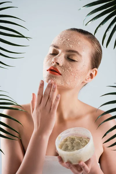 Giovane donna che applica scrub e tiene il contenitore vicino alle foglie di palma sul bianco — Foto stock