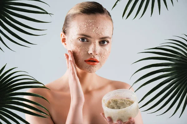 Atractiva mujer joven aplicando exfoliante y sosteniendo contenedor cerca de hojas de palma en blanco - foto de stock