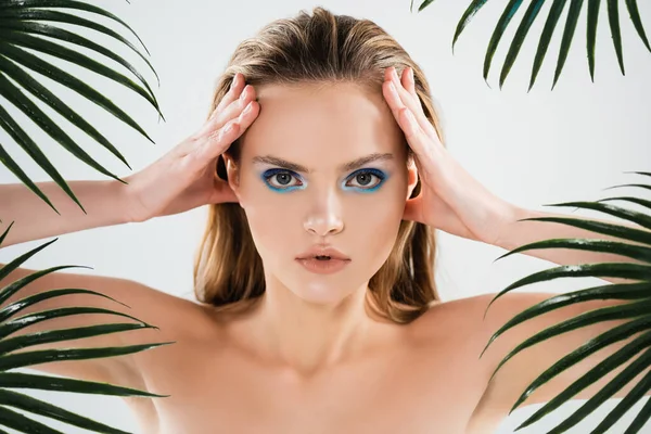 Beautiful woman with blue eye shadow looking at camera and touching face near palm leaves on white — Stock Photo