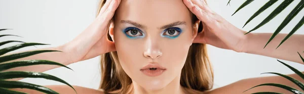 Horizontal crop of beautiful woman with blue eye shadow looking at camera near palm leaves on white — Stock Photo