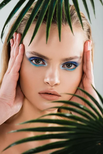 Selective focus of beautiful woman with with blue eye shadow touching face near palm leaves on white — Stock Photo