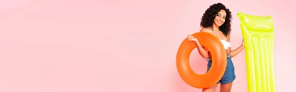 Panoramic crop of cheerful african american girl holding swim ring and inflatable mattress isolated on pink — Stock Photo