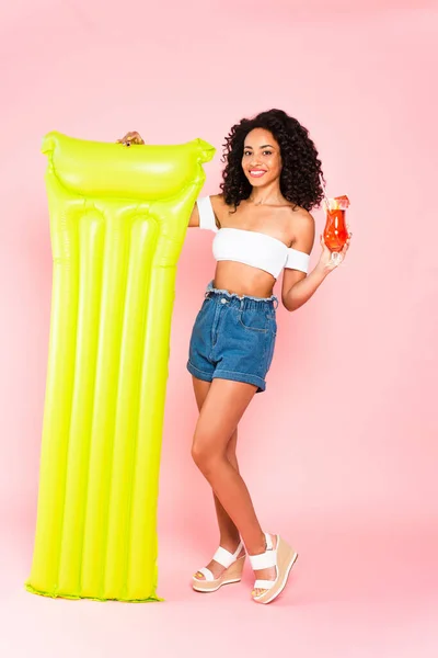 Happy african american girl holding cocktail and inflatable mattress on pink — Stock Photo