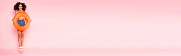 Panoramic shot of excited african american woman holding swim ring and jumping on pink — Stock Photo
