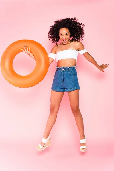 Excitada mujer afroamericana sosteniendo anillo de natación y saltando sobre rosa - foto de stock