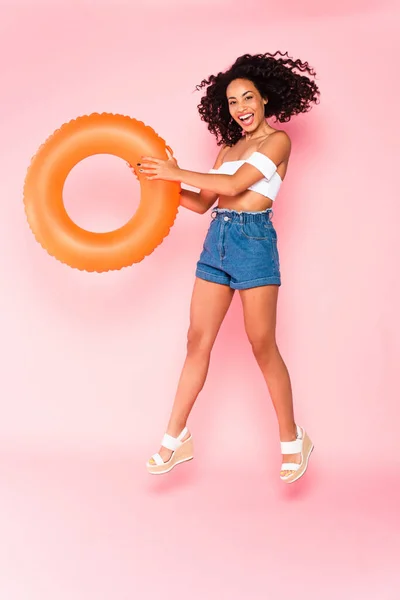 Cheerful african american girl holding swim ring and jumping on pink — Stock Photo