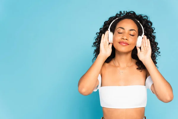 Chica americana con los ojos cerrados escuchando música y tocando auriculares inalámbricos aislados en azul — Stock Photo