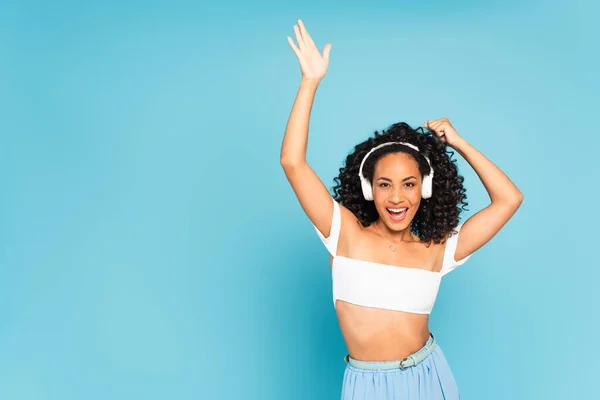 Chica afroamericana emocionada escuchando música en auriculares inalámbricos y bailando en azul - foto de stock
