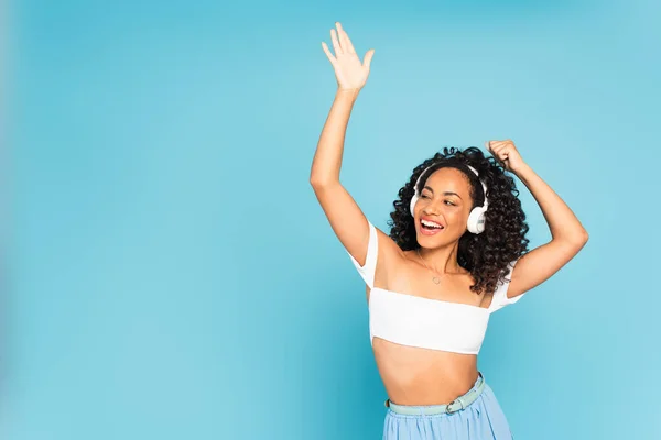 Excited african american woman listening music in wireless headphones and dancing on blue — Stock Photo