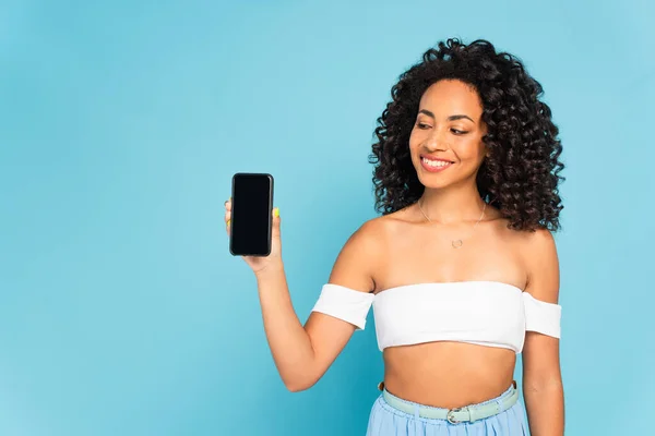 Улыбающаяся африканская американка, выглядящая как смартфон с пустым экраном, изолированным на синем — стоковое фото