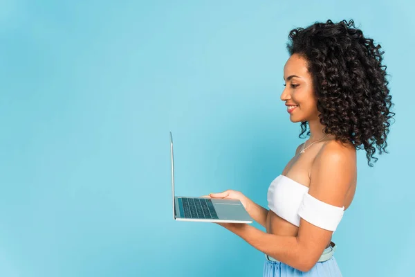 Profile of happy and curly african american woman using laptop isolated on blue — Stock Photo