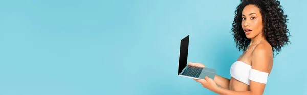 Panoramic concept of surprised and curly african american woman holding laptop isolated on blue — Stock Photo