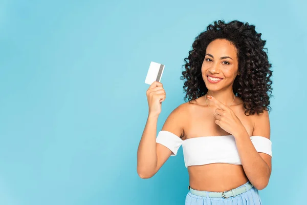 Curly african american woman pointing with finger at credit card isolated on blue — Stock Photo