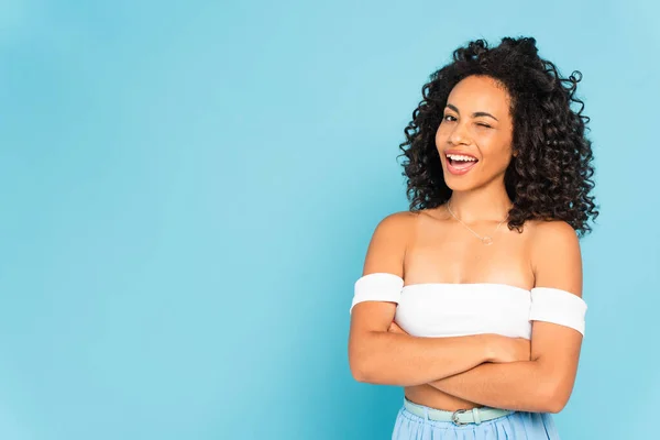 Alegre afro-americana mulher piscando olho e de pé com braços cruzados isolado no azul — Fotografia de Stock