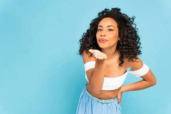 Alegre afroamericana mujer de pie con la mano en la cadera y el envío de aire beso aislado en azul - foto de stock