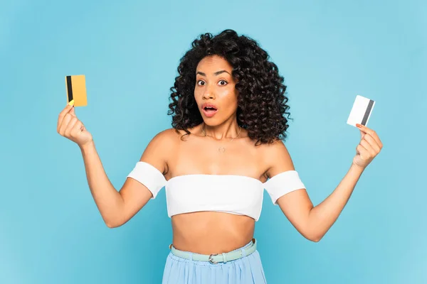 Mulher afro-americana surpreendida segurando cartões de crédito isolados em azul — Fotografia de Stock