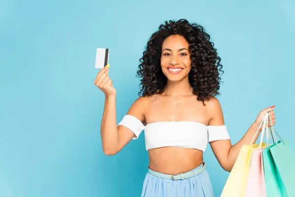 Mulher americana africana alegre segurando cartão de crédito e sacos de compras isolados em azul — Fotografia de Stock