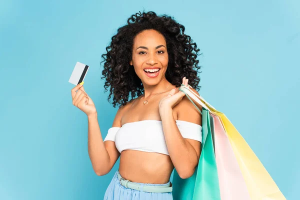 Animado afro-americano mulher segurando cartão de crédito e sacos de compras isolados em azul — Fotografia de Stock