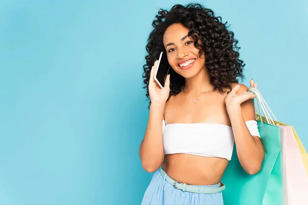 Alegre afroamericana mujer sosteniendo bolsas de compras y hablando en teléfono inteligente aislado en azul - foto de stock