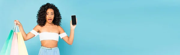 Foto panorámica de la mujer afroamericana sorprendida sosteniendo bolsas de compras y teléfono inteligente con pantalla en blanco aislado en azul - foto de stock