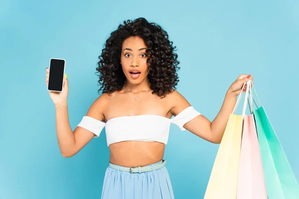 Surpris femme afro-américaine tenant des sacs à provisions et smartphone avec écran blanc isolé sur bleu — Photo de stock