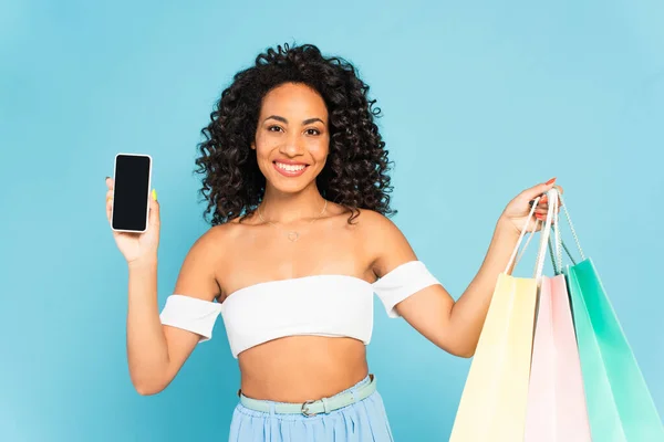 Femme afro-américaine souriante tenant des sacs à provisions et smartphone avec écran blanc isolé sur bleu — Photo de stock