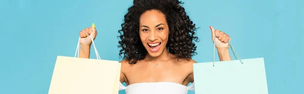 Plano panorámico de la mujer afroamericana emocionada sosteniendo bolsas de compras y sonriendo aislado en azul - foto de stock