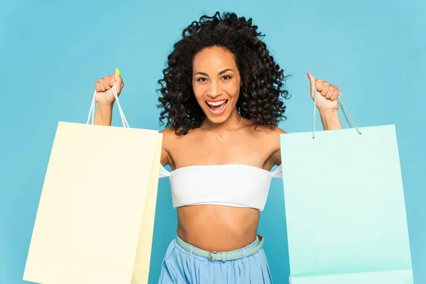 Animado afro-americano mulher segurando sacos de compras e sorrindo isolado no azul — Fotografia de Stock