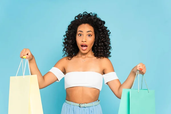 Chocado Africano americano menina com boca aberta segurando sacos de compras isolados em azul — Fotografia de Stock